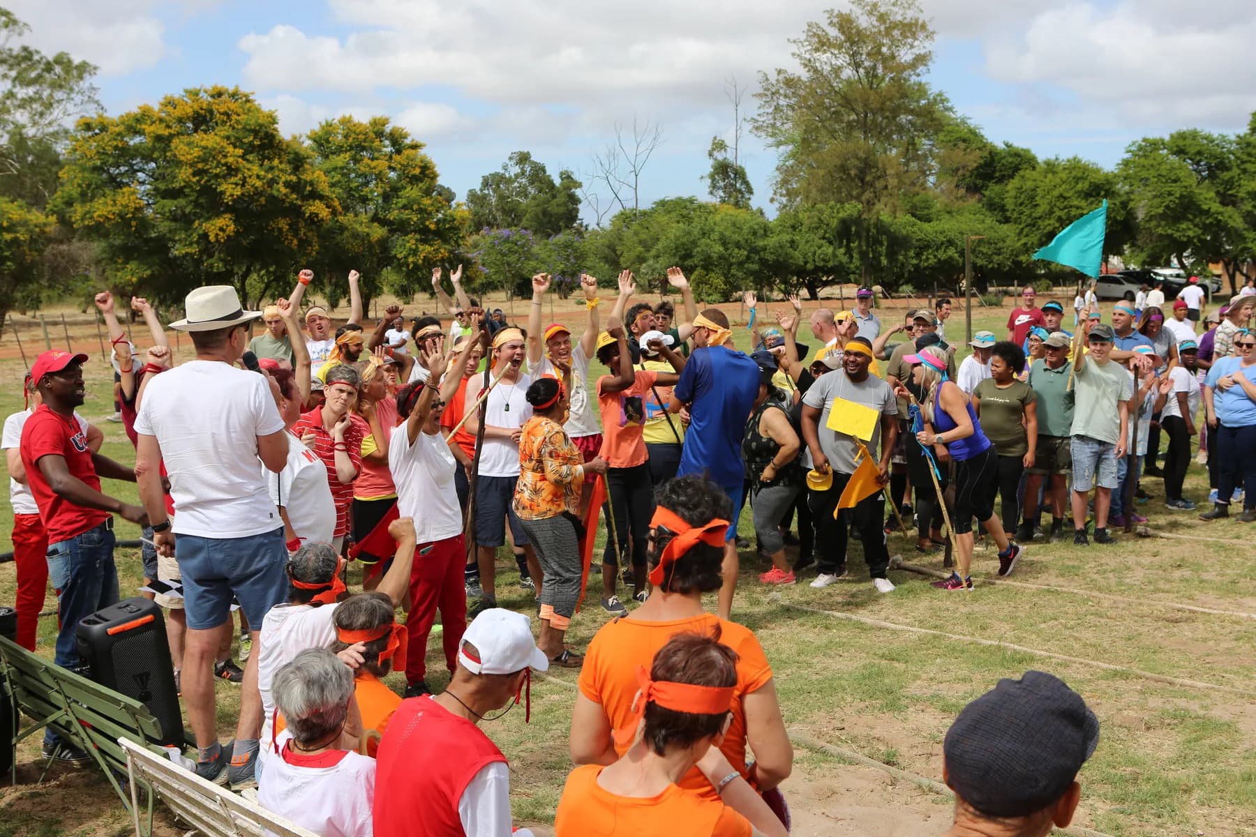A crowd of Camphill Village residents and staff in brightly coloured team bibs and headbands, cheering with their arms in the air at the village sports day.