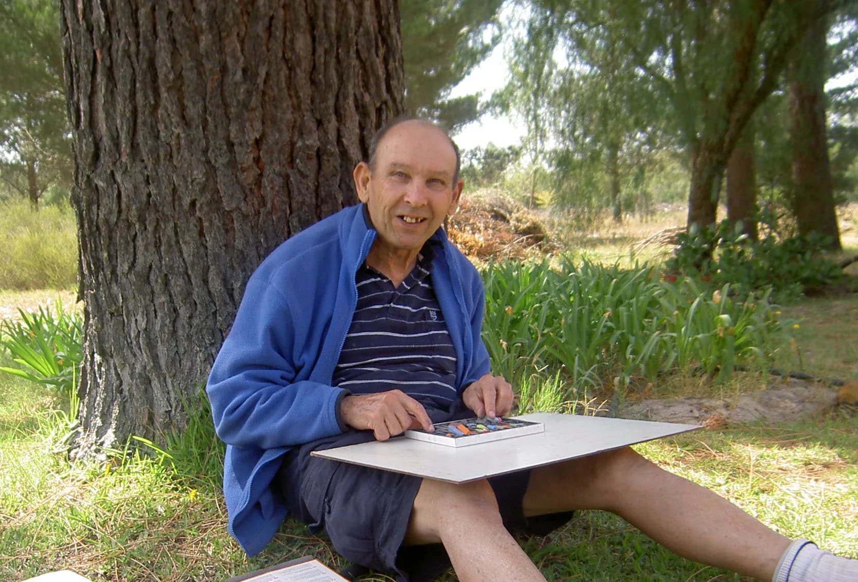 A resident sitting in the shade of a pine tree with a drawing board on his lap and a tin of pastels open beside him.